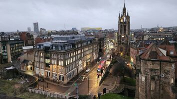 A view of a Newcastle city street in the early evening with historic buildings, buses, and a prominent church tower in the background, under a cloudy sky