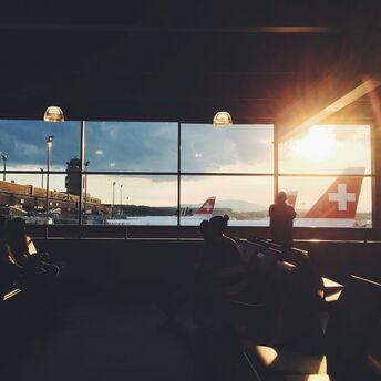 Passengers waiting in an airport lounge with Swiss Air planes visible through the large windows as the sun sets