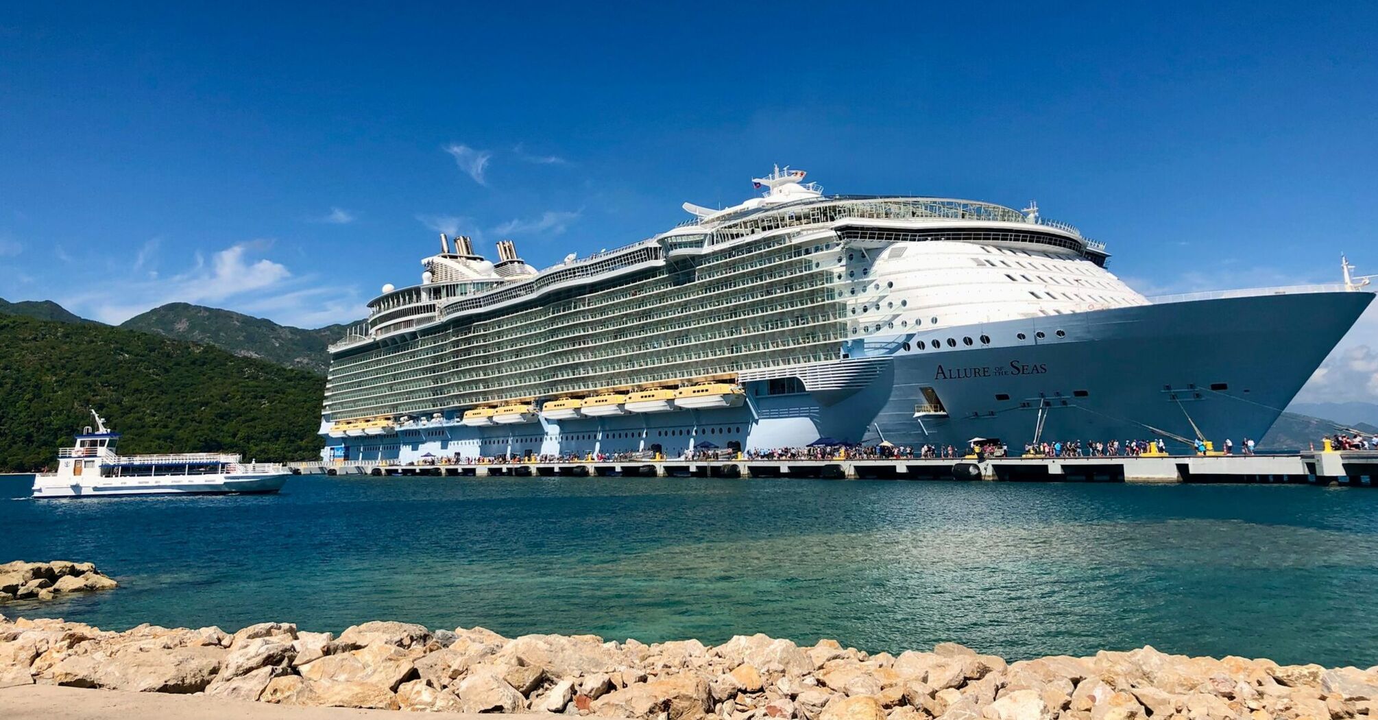 A large Royal Caribbean cruise ship docked at a scenic port with a smaller boat nearby and lush green hills in the background under a clear blue sky