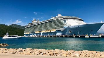 A large Royal Caribbean cruise ship docked at a scenic port with a smaller boat nearby and lush green hills in the background under a clear blue sky