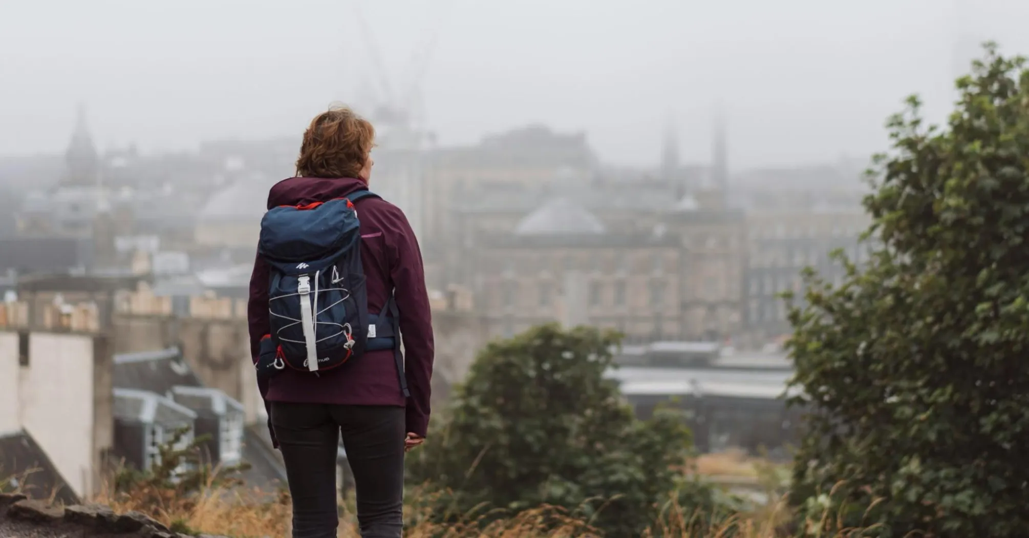 A person with a backpack stands on a hill, overlooking the misty skyline of Edinburgh