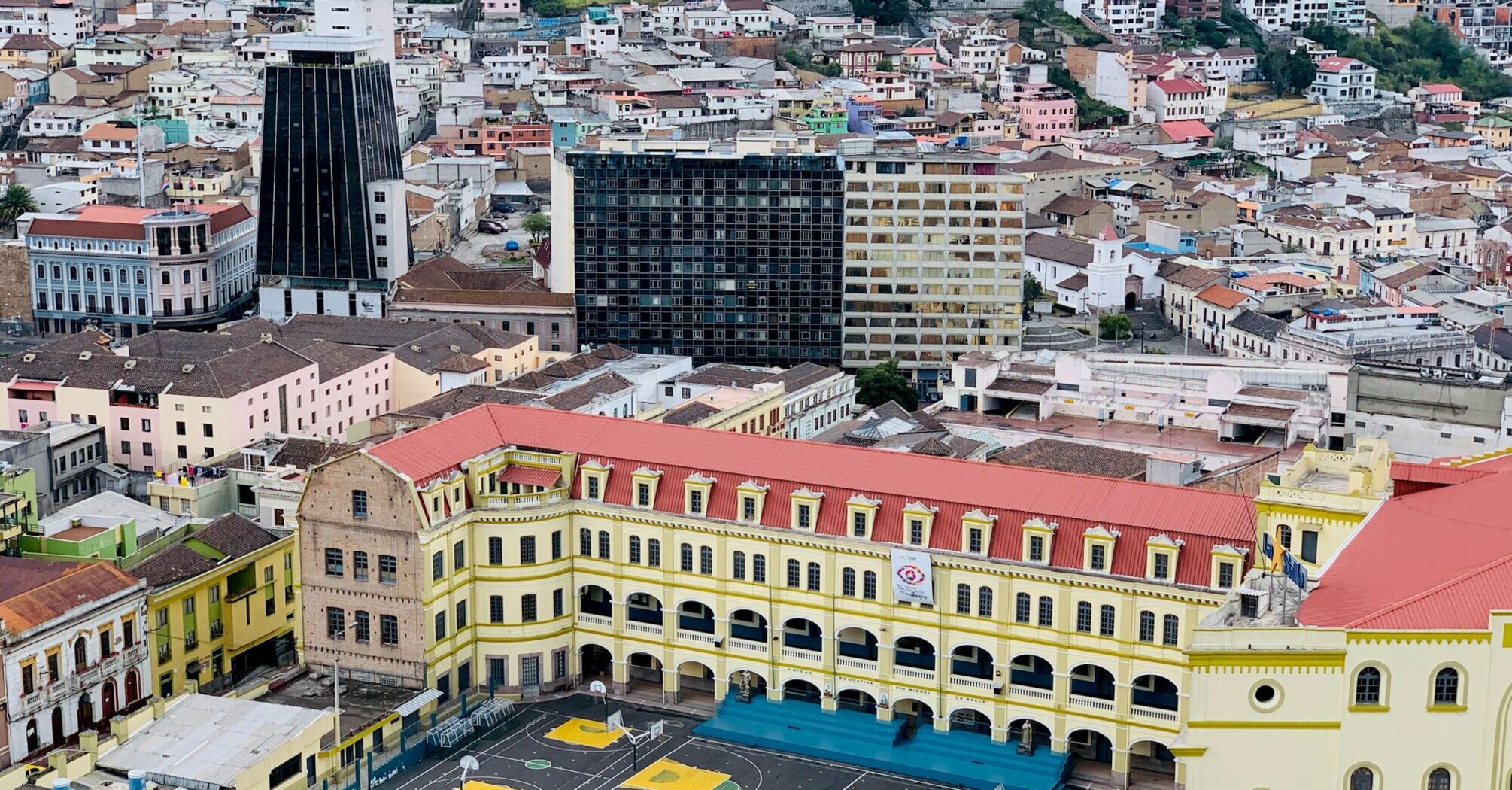 Aerial view of a historic yellow building with a red roof in Quito, Ecuador, surrounded by colorful urban buildings. The foreground shows a sports court with yellow markings