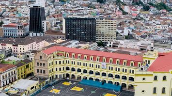 Aerial view of a historic yellow building with a red roof in Quito, Ecuador, surrounded by colorful urban buildings. The foreground shows a sports court with yellow markings