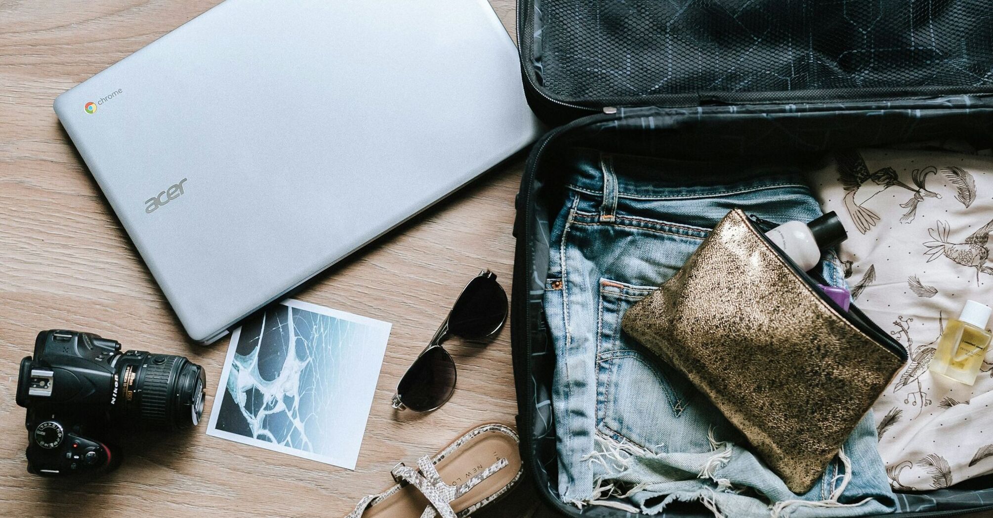 A packed suitcase with travel essentials including a laptop, camera, sunglasses, sandals, and toiletries laid out on a wooden floor