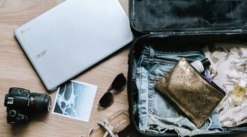 A packed suitcase with travel essentials including a laptop, camera, sunglasses, sandals, and toiletries laid out on a wooden floor