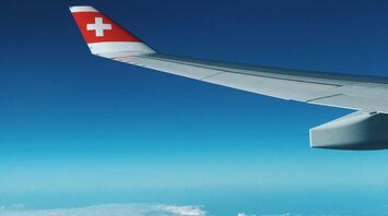 View of airplane wing with Swiss flag flying above the clouds and mountains