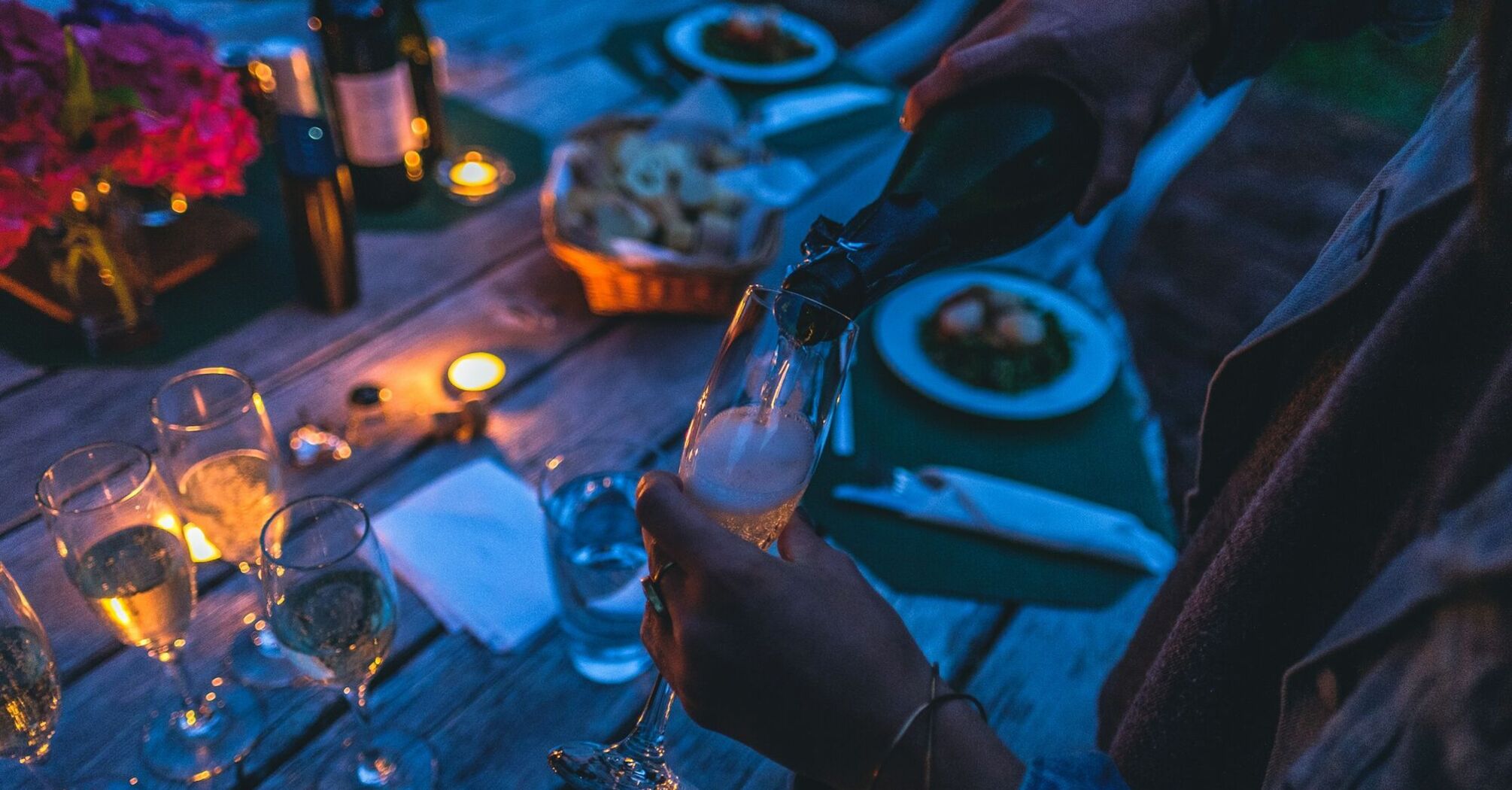 A person pouring champagne into a glass at a candlelit outdoor dinner setting with food and wine on the table