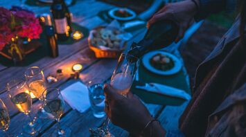 A person pouring champagne into a glass at a candlelit outdoor dinner setting with food and wine on the table