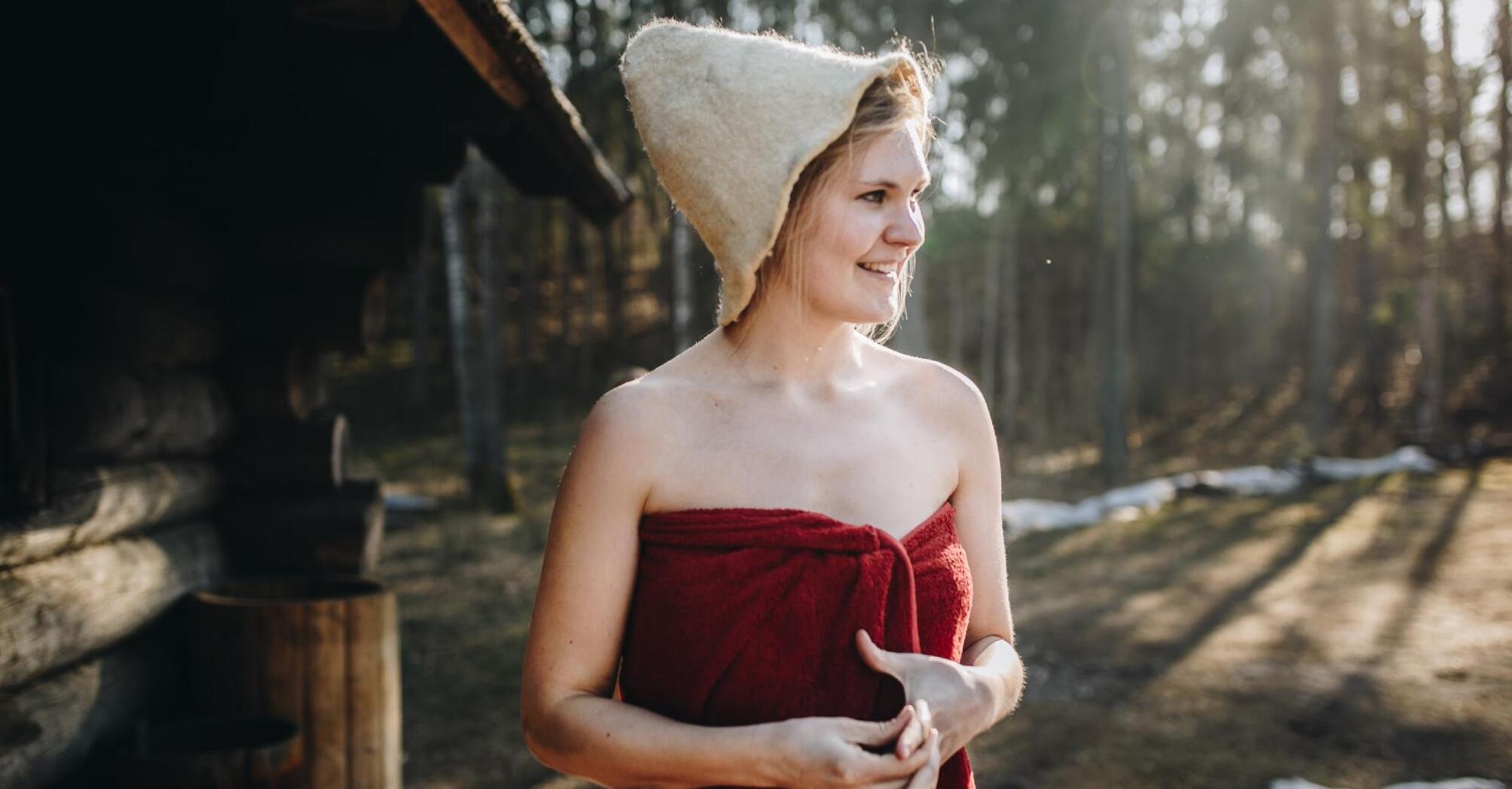 A woman in a traditional felt sauna hat, wrapped in a red towel, smiling outdoors near a wooden structure