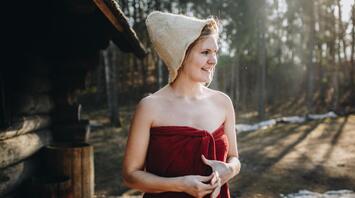 A woman in a traditional felt sauna hat, wrapped in a red towel, smiling outdoors near a wooden structure