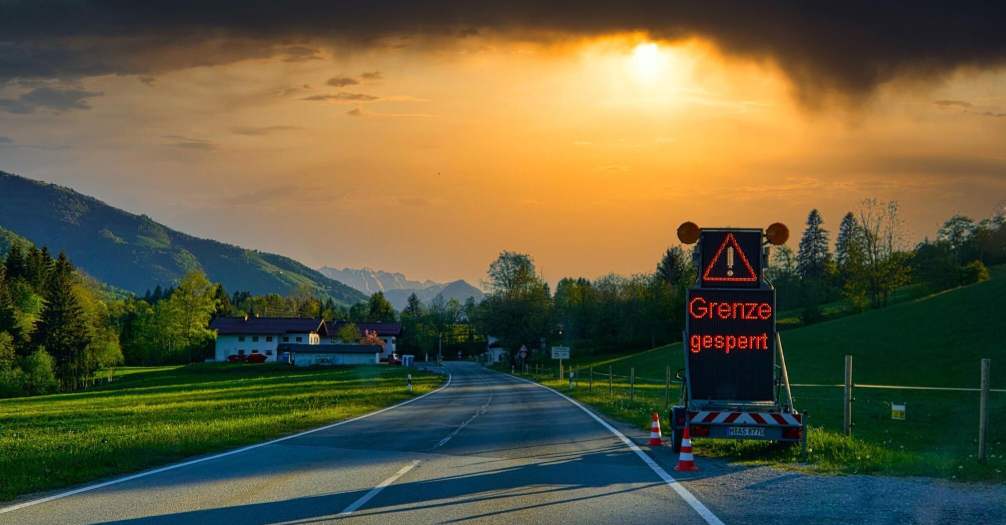 Road leading to a sunset, with a "Grenze gesperrt" (border closed) sign prominently displayed, set against a backdrop of lush green fields and distant mountains