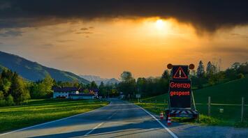 Road leading to a sunset, with a "Grenze gesperrt" (border closed) sign prominently displayed, set against a backdrop of lush green fields and distant mountains