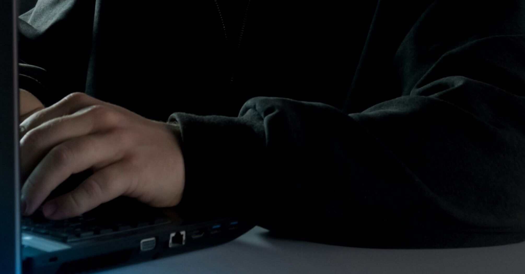 A hooded figure using a laptop in a dark setting, symbolizing online fraud or cybercrime