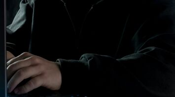 A hooded figure using a laptop in a dark setting, symbolizing online fraud or cybercrime