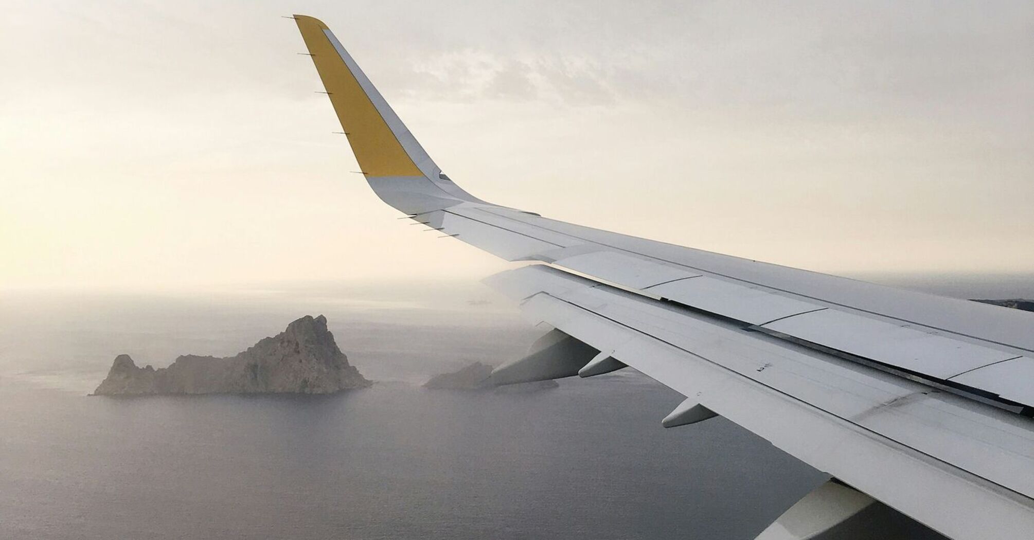 View from airplane window showing a distant island over the sea, with a plane wing in the foreground under a cloudy sky