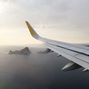 View from airplane window showing a distant island over the sea, with a plane wing in the foreground under a cloudy sky