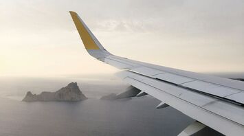 View from airplane window showing a distant island over the sea, with a plane wing in the foreground under a cloudy sky