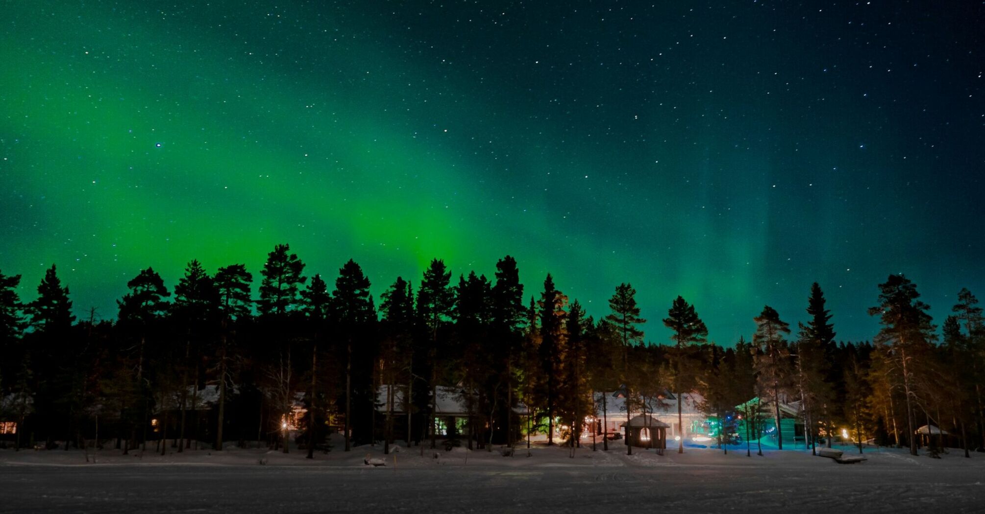 Northern lights illuminating the sky over a snowy forest with cabins below