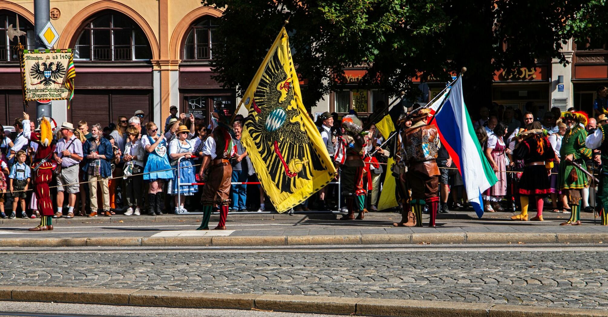 A traditional Bavarian parade during Oktoberfest featuring participants in historic costumes, holding colorful flags, while spectators in traditional attire watch from the sidewalk