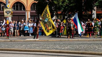 A traditional Bavarian parade during Oktoberfest featuring participants in historic costumes, holding colorful flags, while spectators in traditional attire watch from the sidewalk