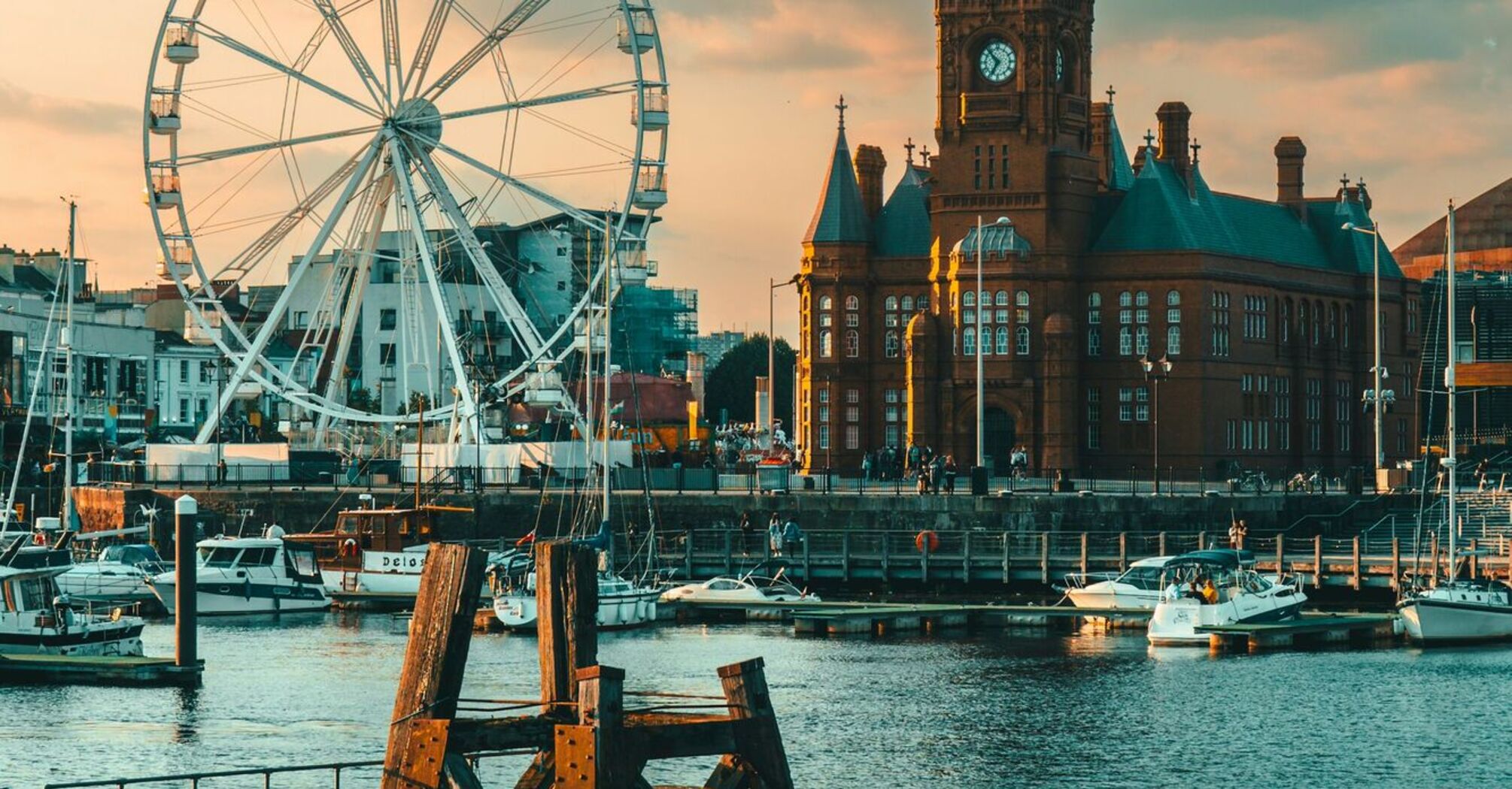 A view of Cardiff Bay featuring the Pierhead Building and a Ferris wheel at sunset, with boats docked in the harbor