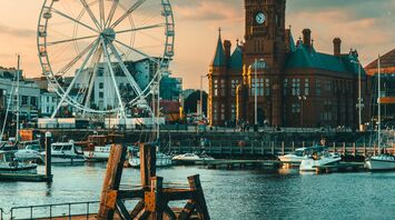 A view of Cardiff Bay featuring the Pierhead Building and a Ferris wheel at sunset, with boats docked in the harbor