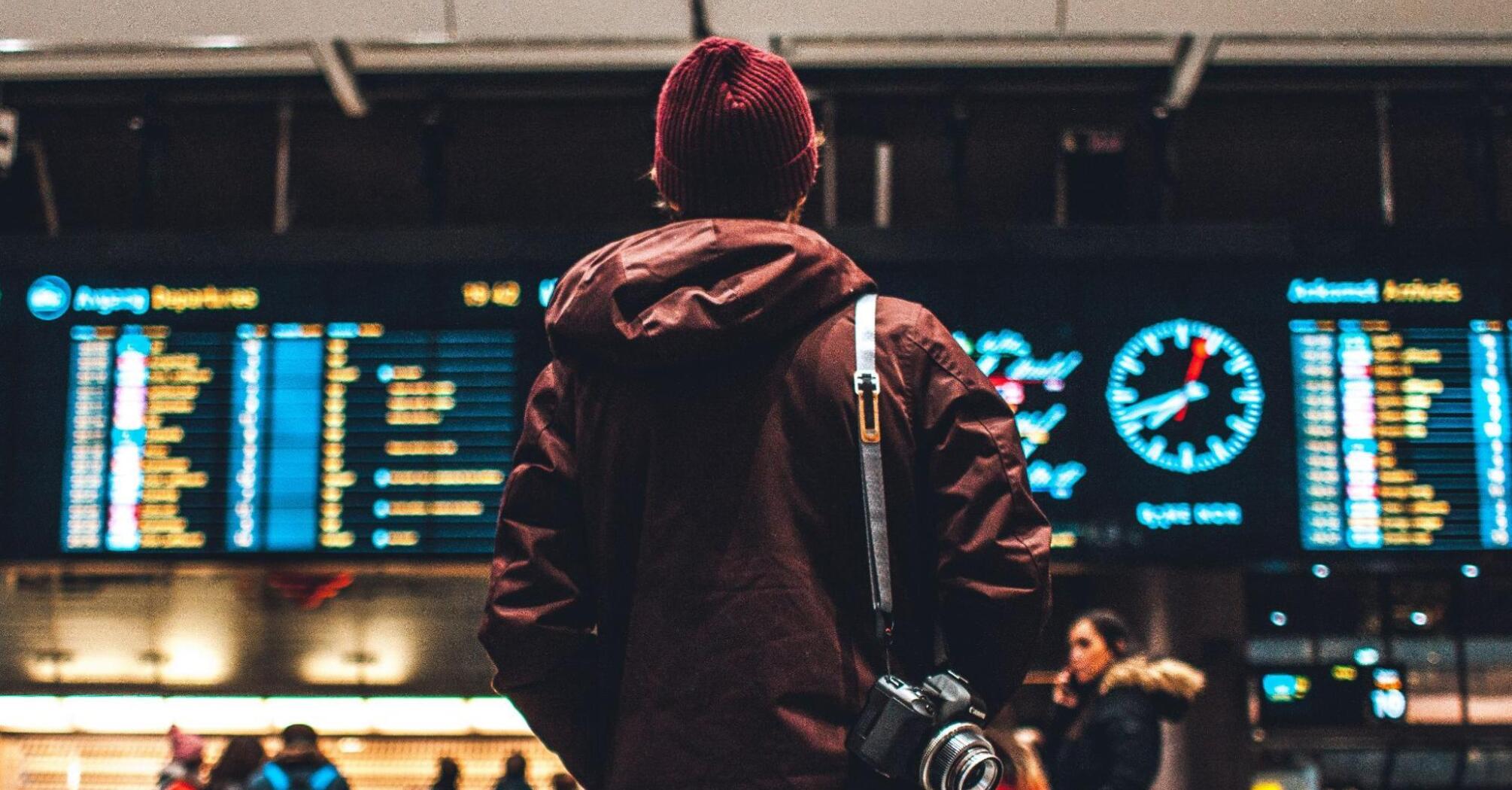 A traveler with a camera around their neck looks up at an airport departure board, contemplating their next destination