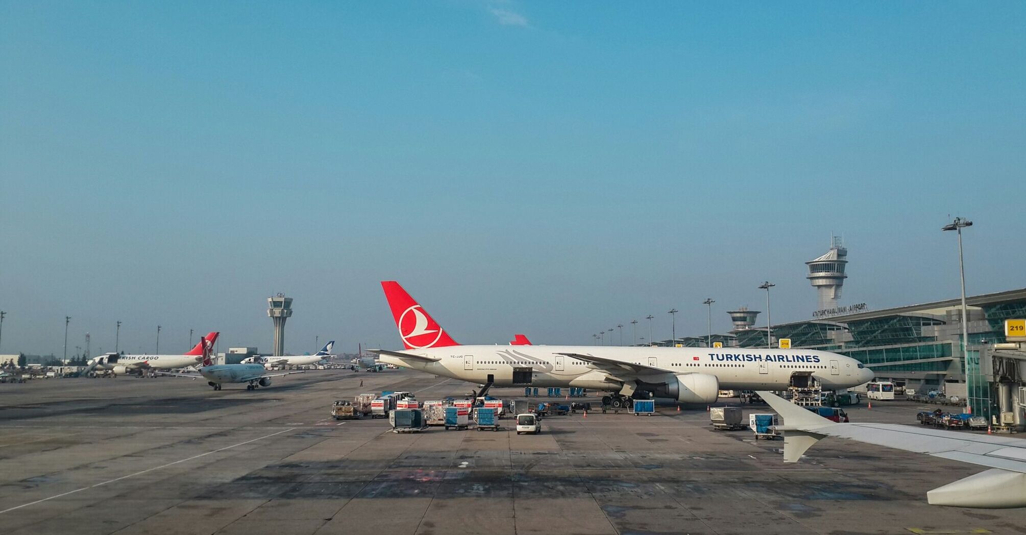 Turkish Airlines aircraft at an international airport gate, with cargo being loaded and airport control towers in the background