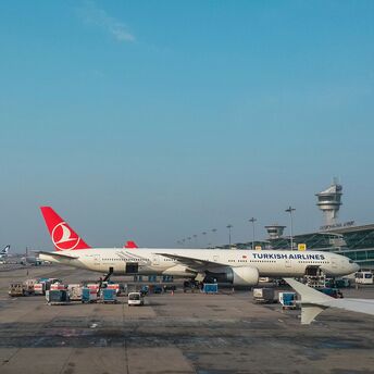 Turkish Airlines aircraft at an international airport gate, with cargo being loaded and airport control towers in the background
