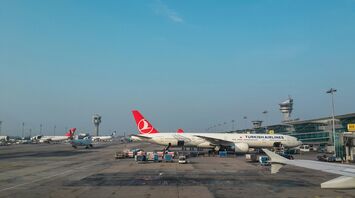 Turkish Airlines aircraft at an international airport gate, with cargo being loaded and airport control towers in the background