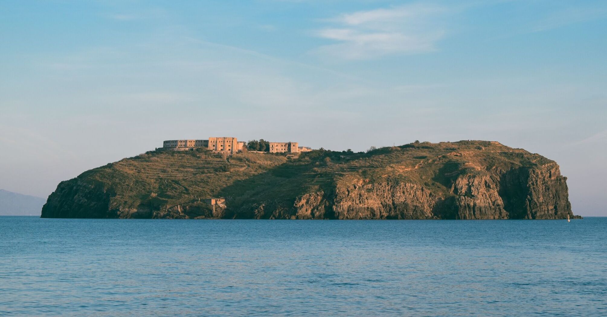 A view of the island of Ventotene with a historic structure perched on its rocky landscape, surrounded by tranquil waters and a bright blue sky