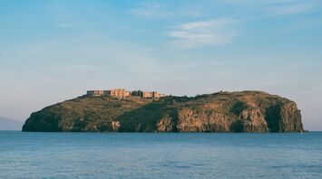 A view of the island of Ventotene with a historic structure perched on its rocky landscape, surrounded by tranquil waters and a bright blue sky