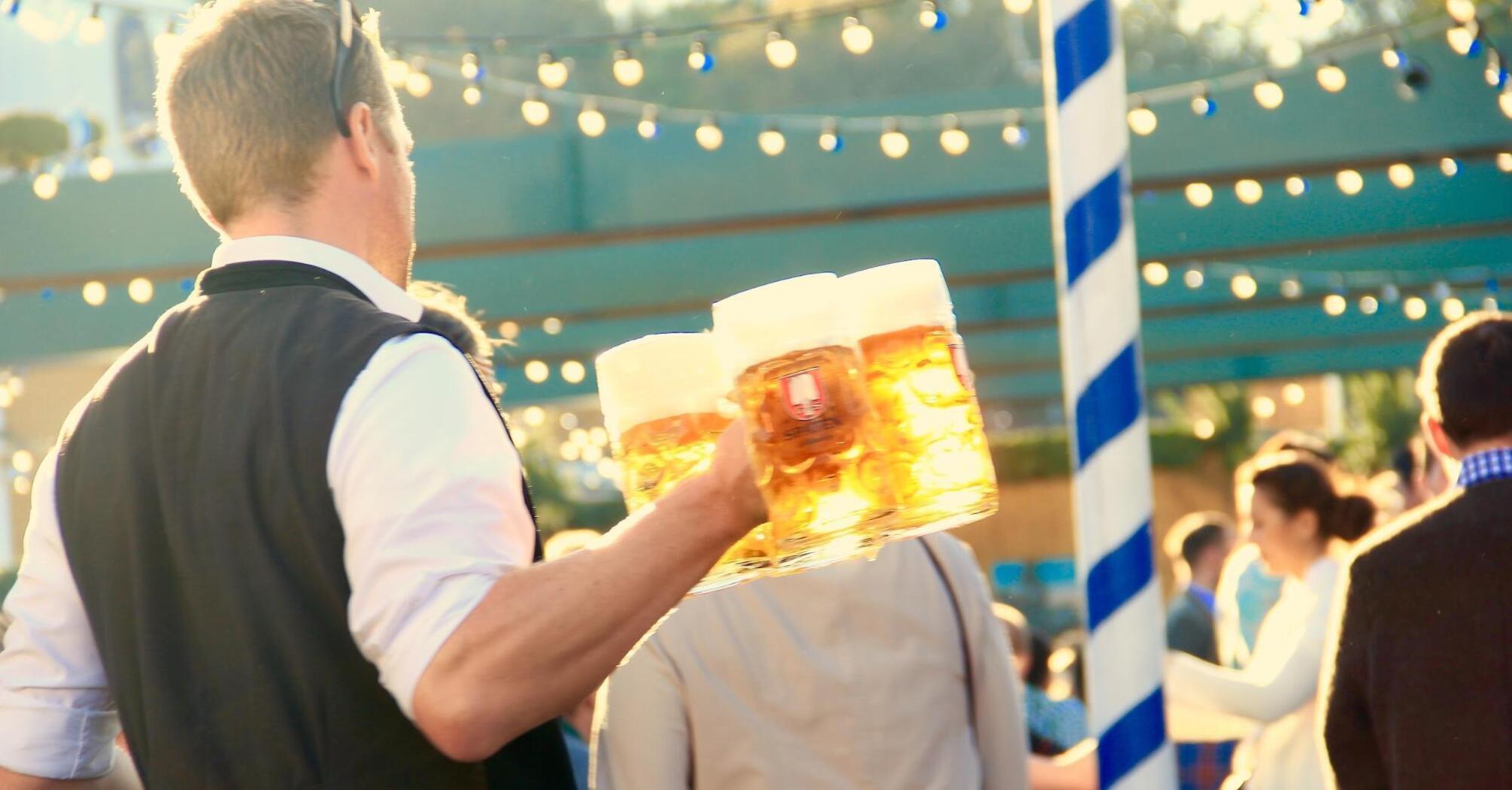 A waiter in traditional attire carrying multiple beer steins at an outdoor festival with string lights