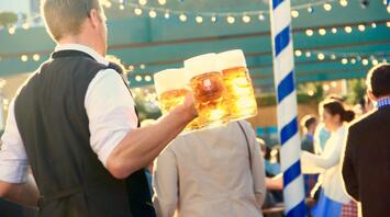 A waiter in traditional attire carrying multiple beer steins at an outdoor festival with string lights