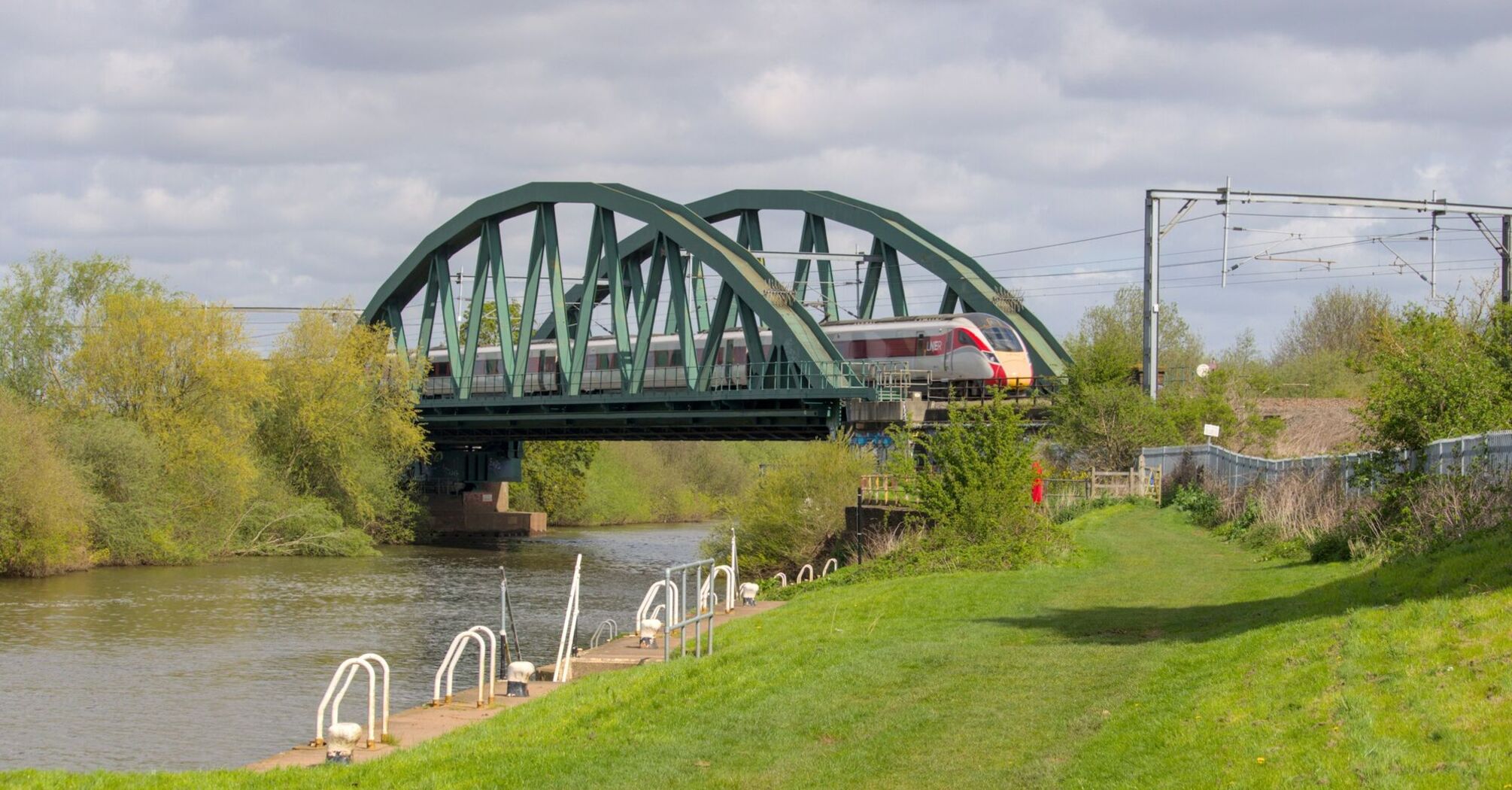 A modern LNER train crossing a green steel bridge over a river, surrounded by trees and greenery