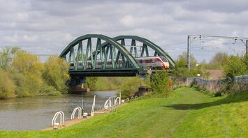 A modern LNER train crossing a green steel bridge over a river, surrounded by trees and greenery