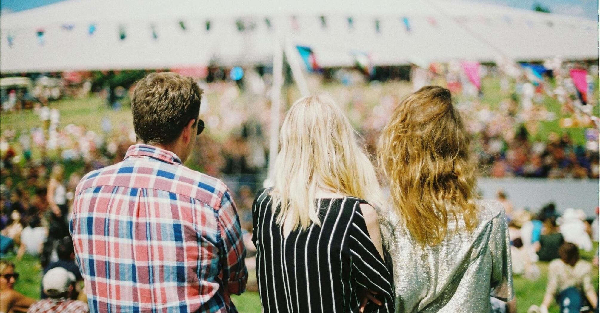 Three people enjoying an outdoor festival with a crowd and a large tent in the background