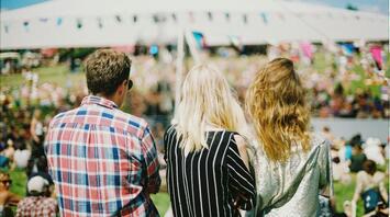 Three people enjoying an outdoor festival with a crowd and a large tent in the background