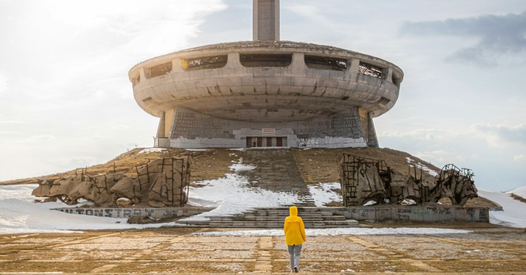 A person in a yellow jacket walks towards the abandoned UFO-shaped Buzludzha Monument in Bulgaria under a cloudy sky