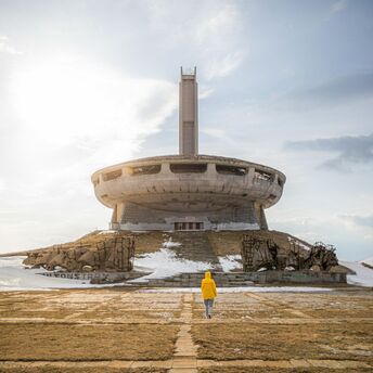 A person in a yellow jacket walks towards the abandoned UFO-shaped Buzludzha Monument in Bulgaria under a cloudy sky