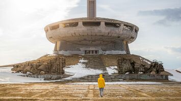 A person in a yellow jacket walks towards the abandoned UFO-shaped Buzludzha Monument in Bulgaria under a cloudy sky