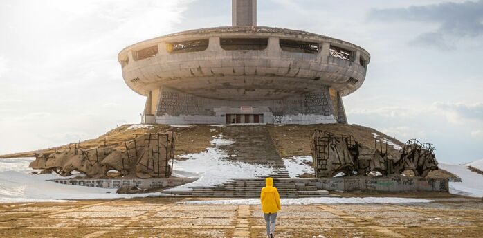 A person in a yellow jacket walks towards the abandoned UFO-shaped Buzludzha Monument in Bulgaria under a cloudy sky