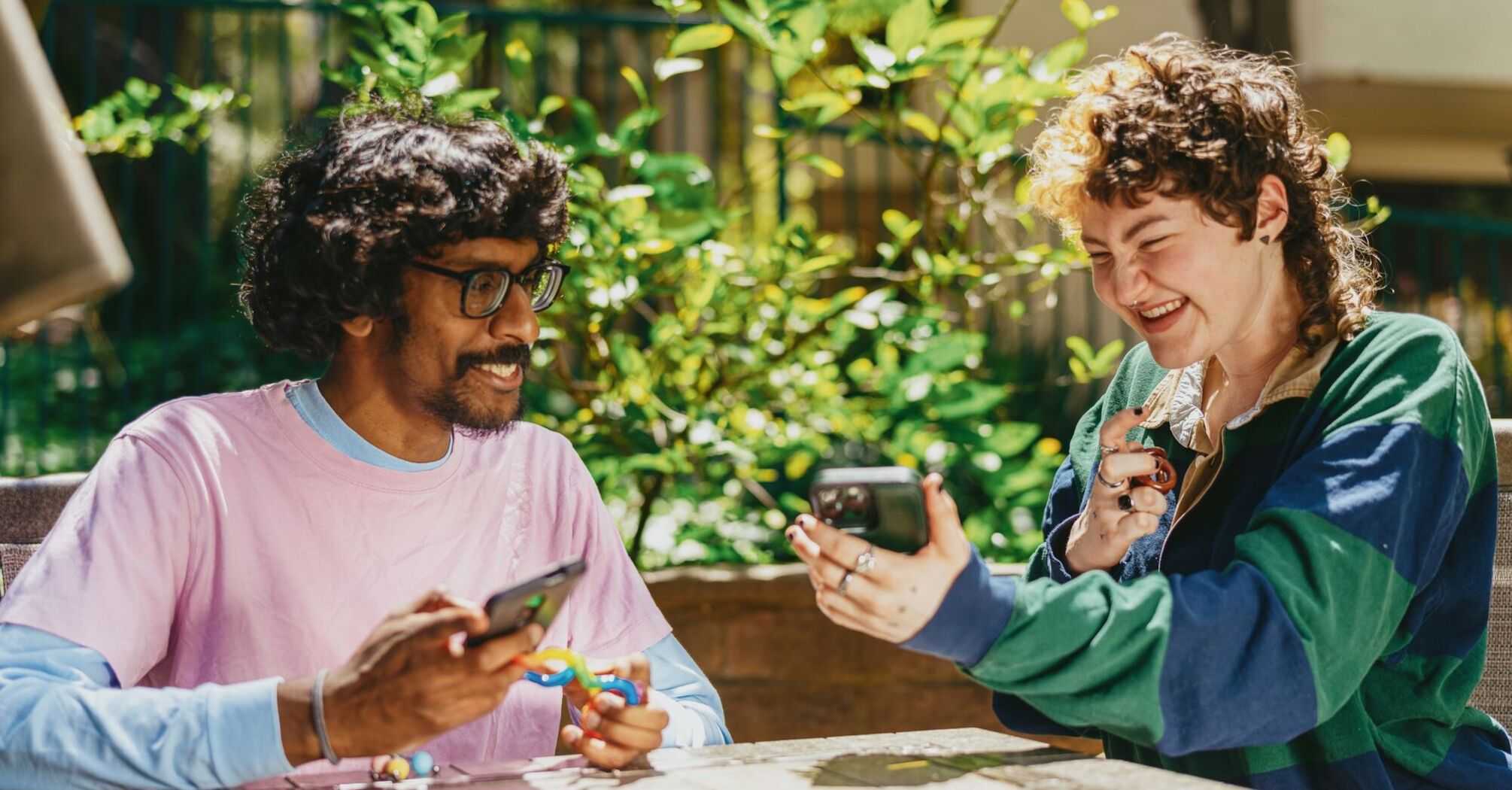 Two friends, including one neurodivergent individual, joyfully sharing content on their smartphones while sitting outdoors, surrounded by greenery