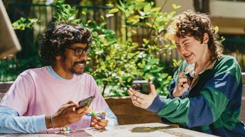 Two friends, including one neurodivergent individual, joyfully sharing content on their smartphones while sitting outdoors, surrounded by greenery