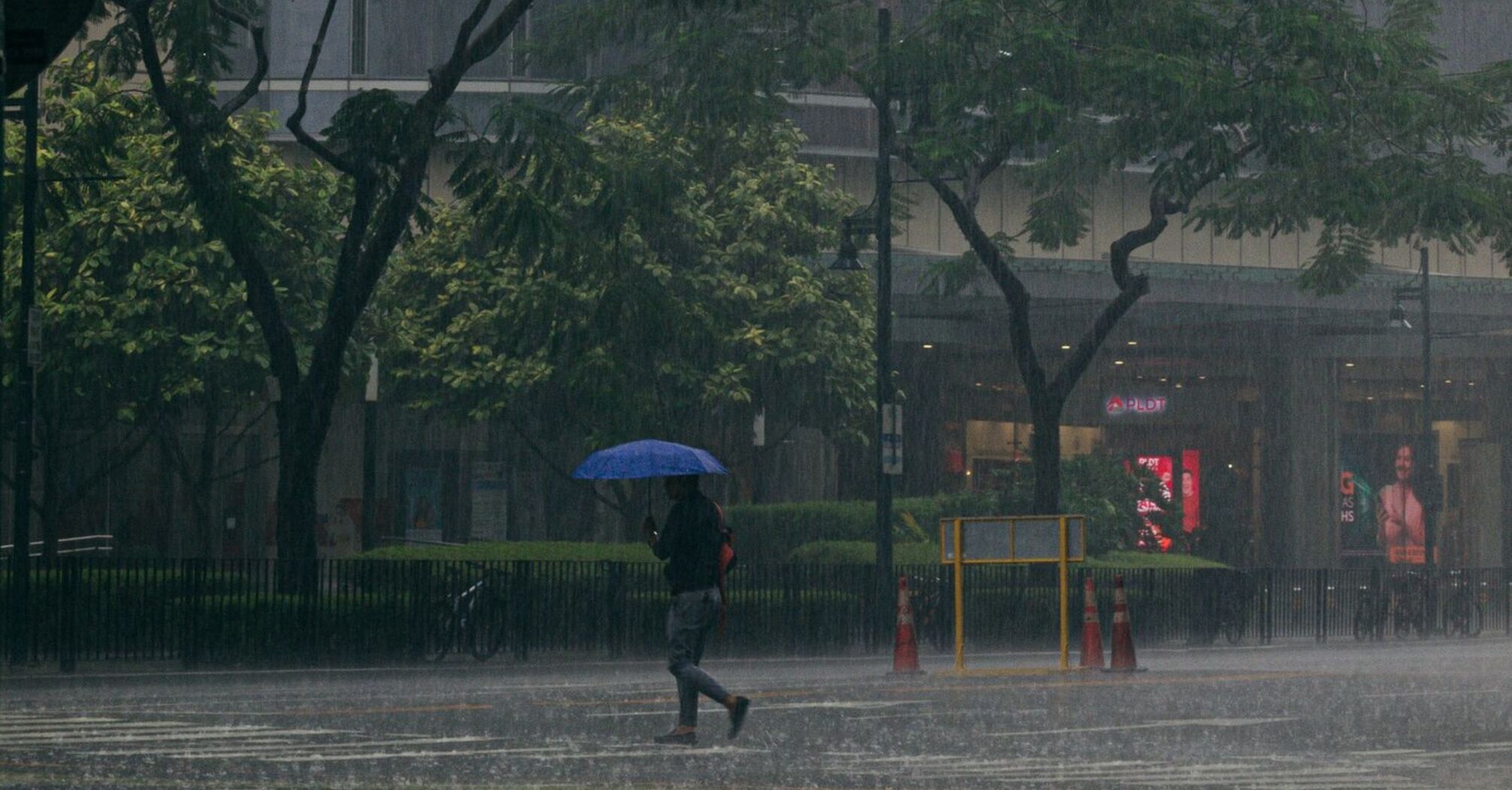 A person holding an umbrella crosses a city street during heavy rainfall, with traffic lights and tall buildings in the background