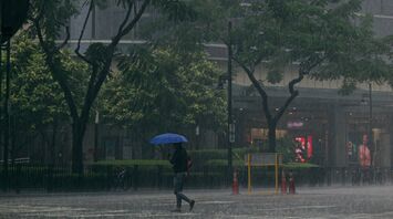 A person holding an umbrella crosses a city street during heavy rainfall, with traffic lights and tall buildings in the background