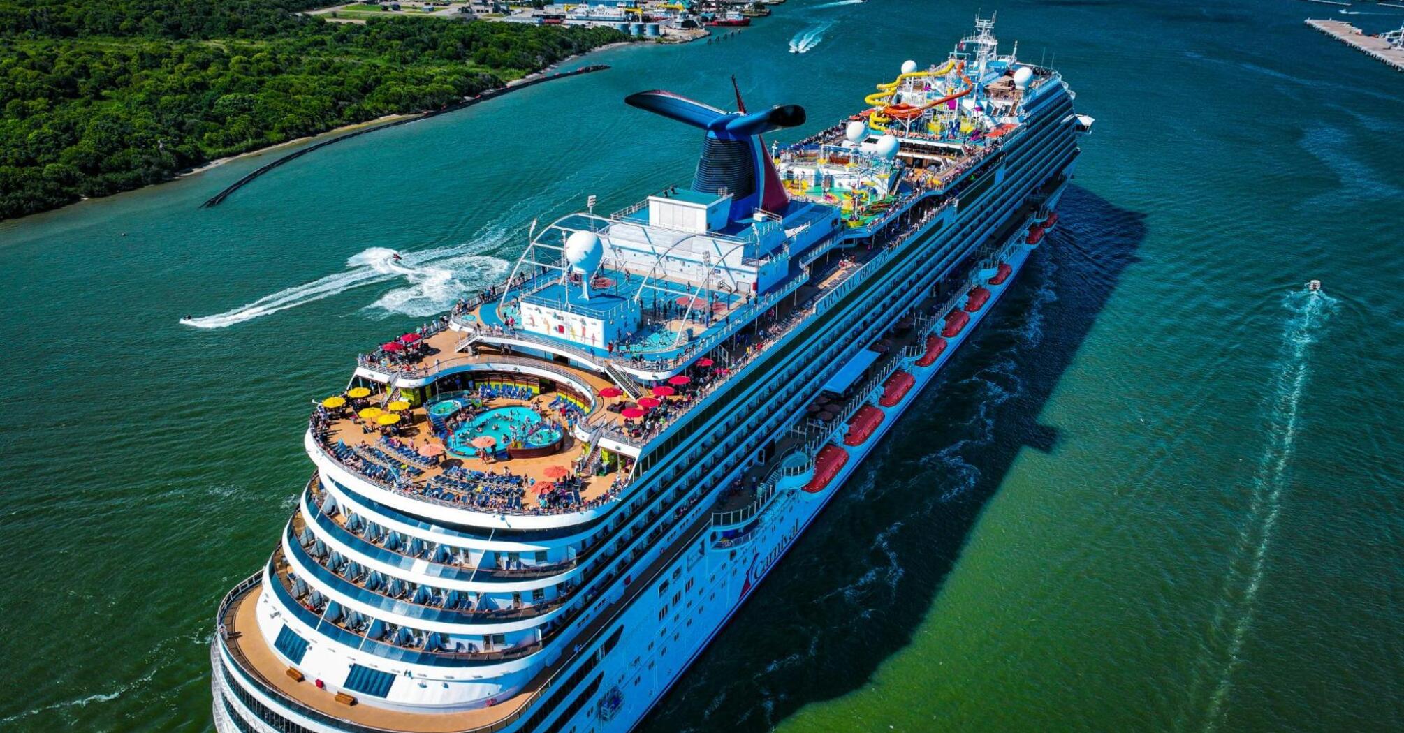 A Carnival cruise ship with a water park and pools on the top deck departs from port with a green coastline in the background