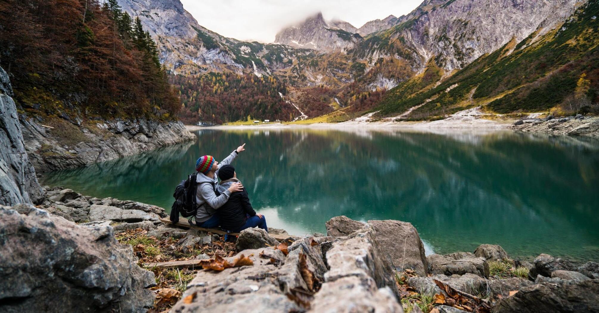 Two hikers pointing at a mountain across a serene lake in autumn