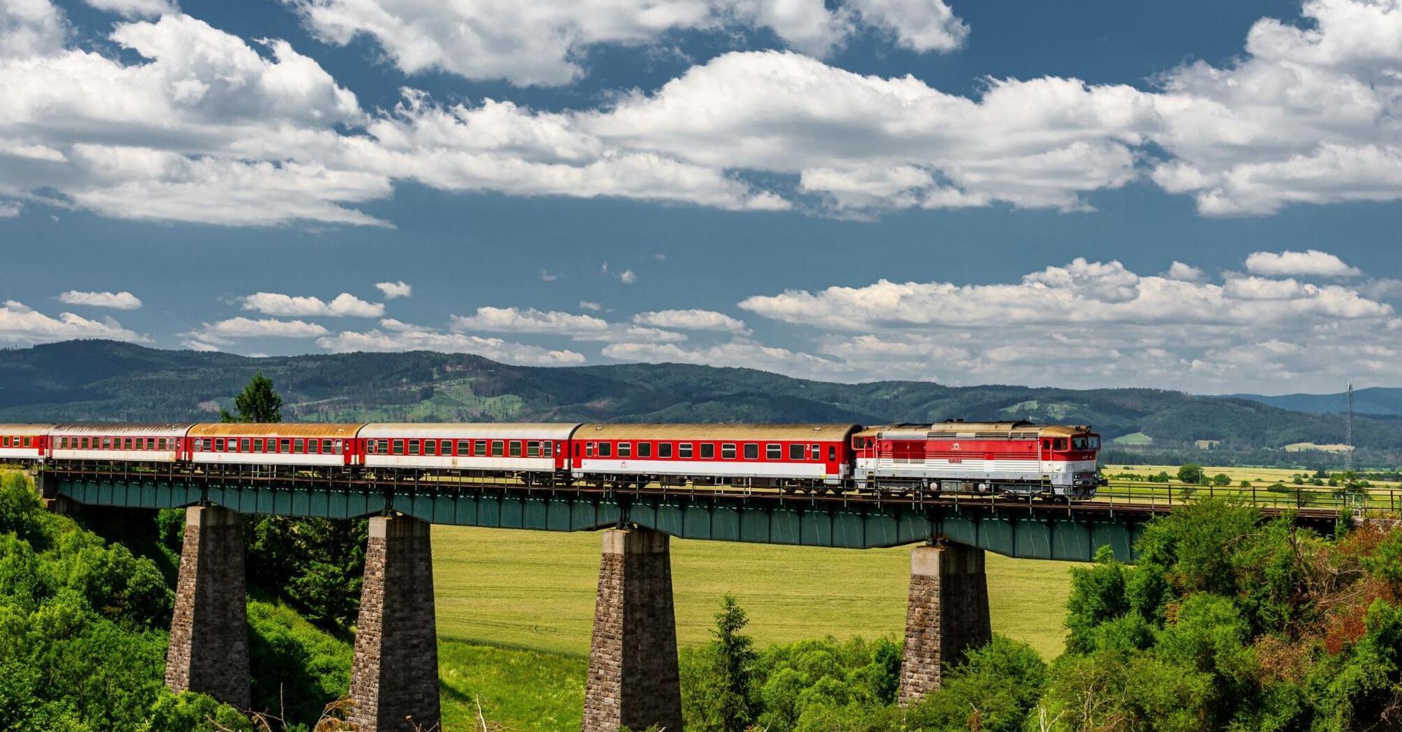 A red passenger train crossing a tall viaduct in a lush, green landscape under a cloudy sky