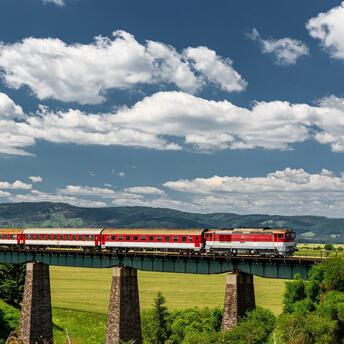 A red passenger train crossing a tall viaduct in a lush, green landscape under a cloudy sky